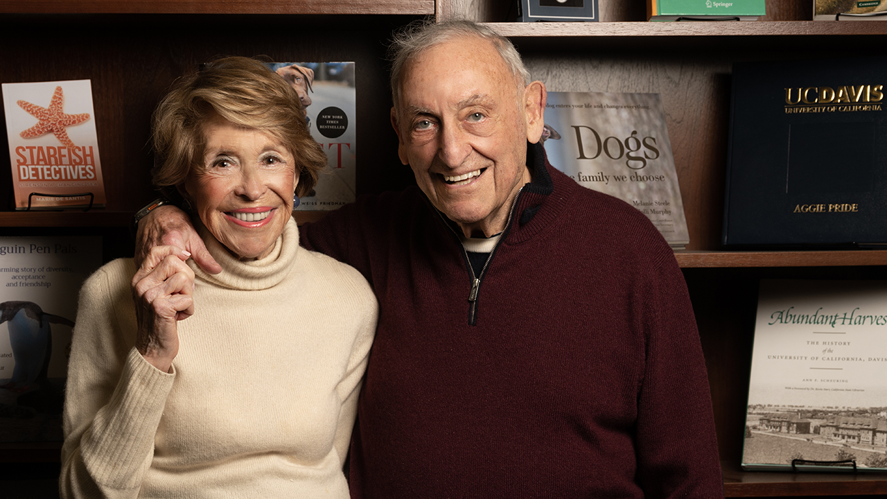 A smiling older couple stands closely together in front of a bookshelf.