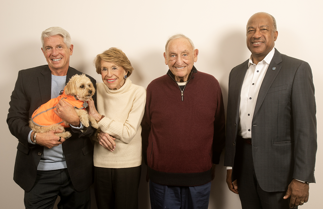 Four people stand together, smiling, with one holding a small dog in an orange vest.