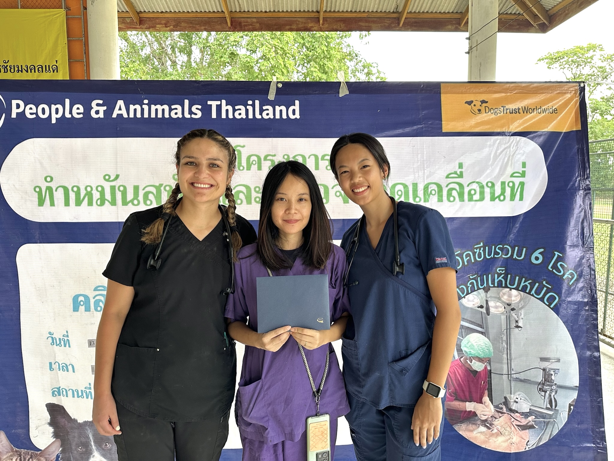 Eliana and 2 other local vets stand in front of a sign with pictures that says "people and animals in Thailand"