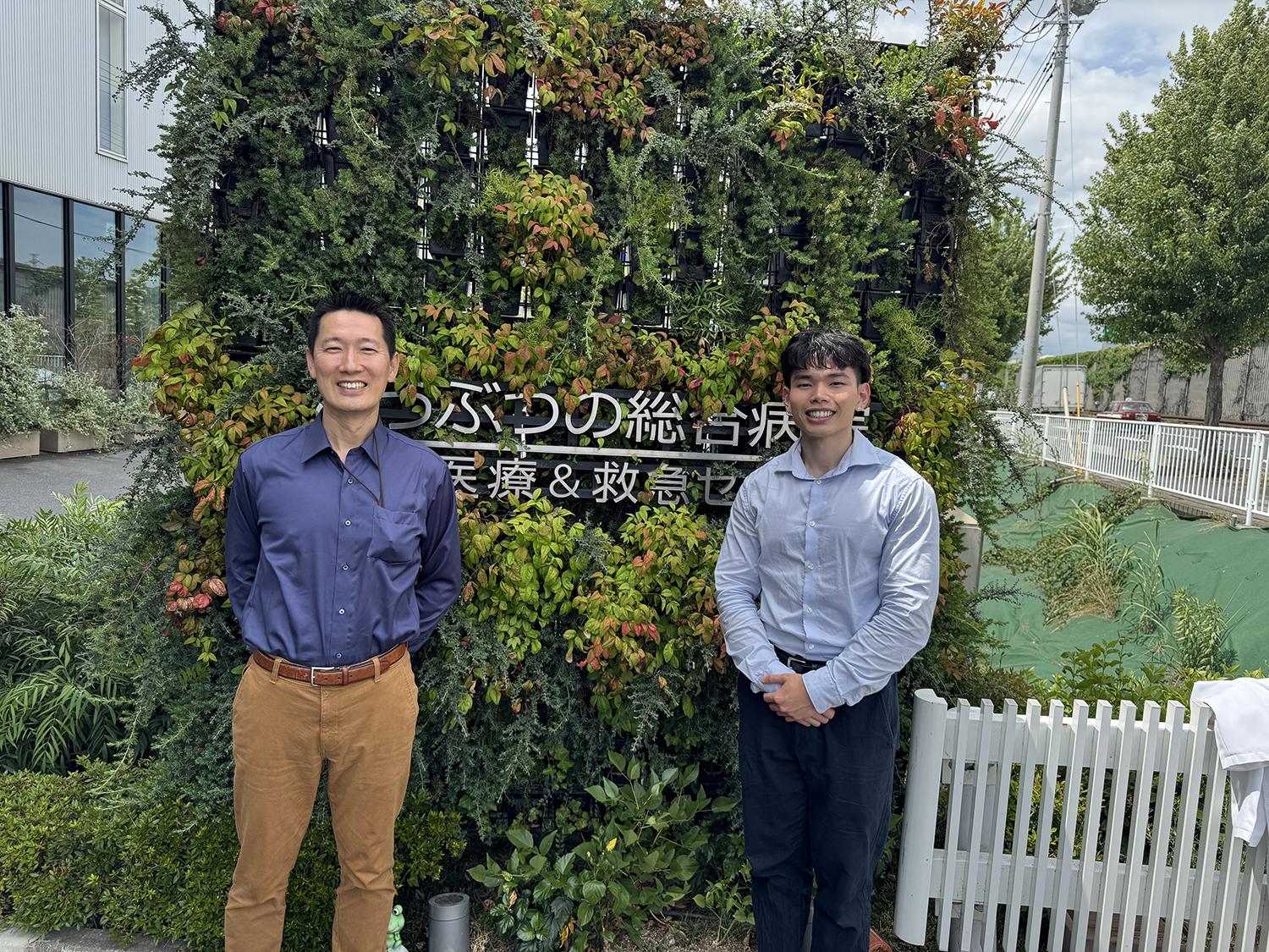 Two people standing in front of an exterior building sign covered in foliage.