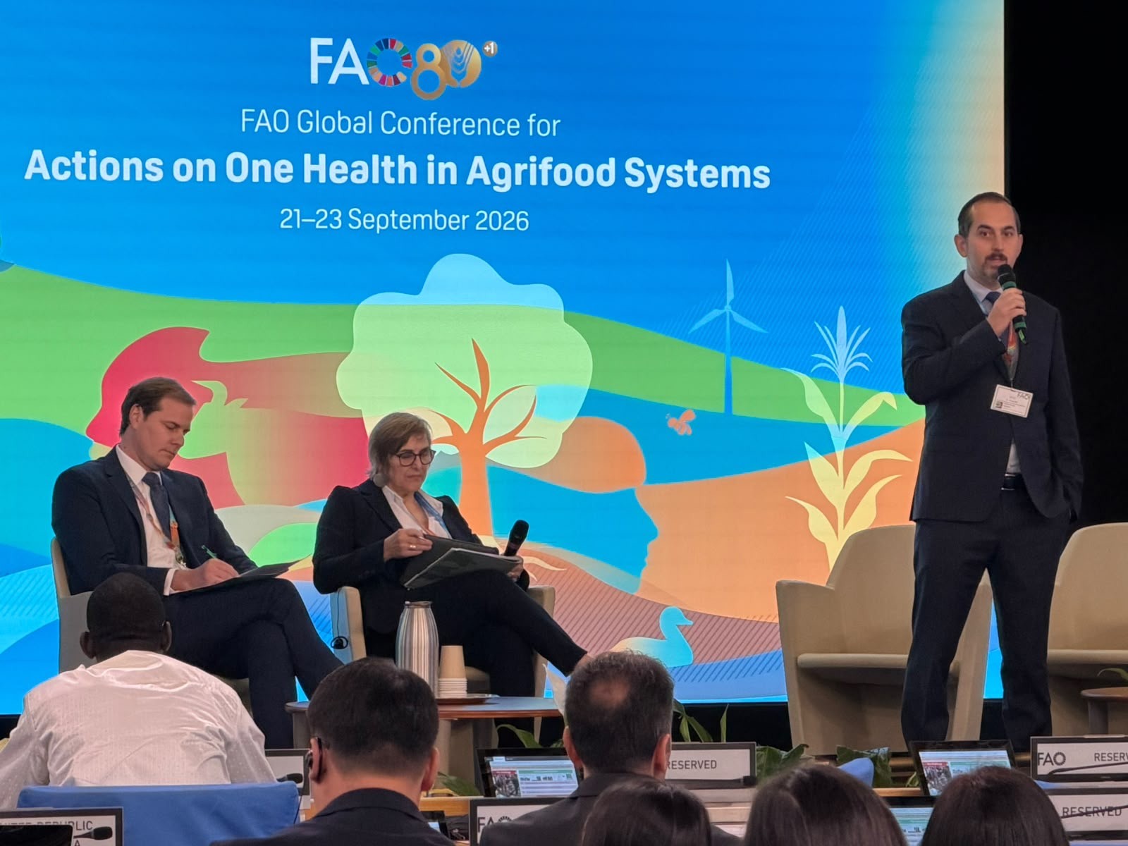 Three presenters on conference stage under blue FAO banner; audience heads in foreground