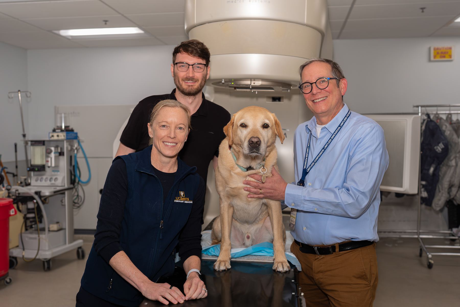Three smiling clinicians and a yellow Labrador on a medical scanner table