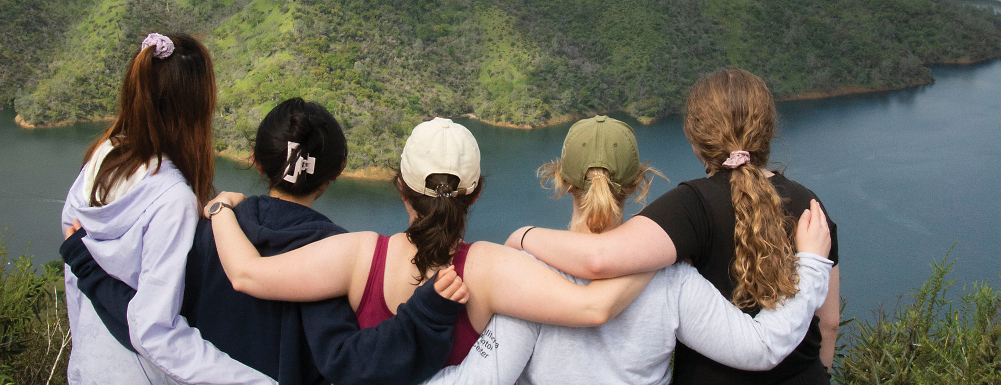 A group of five students on a summit with their arms around each other facing the view.