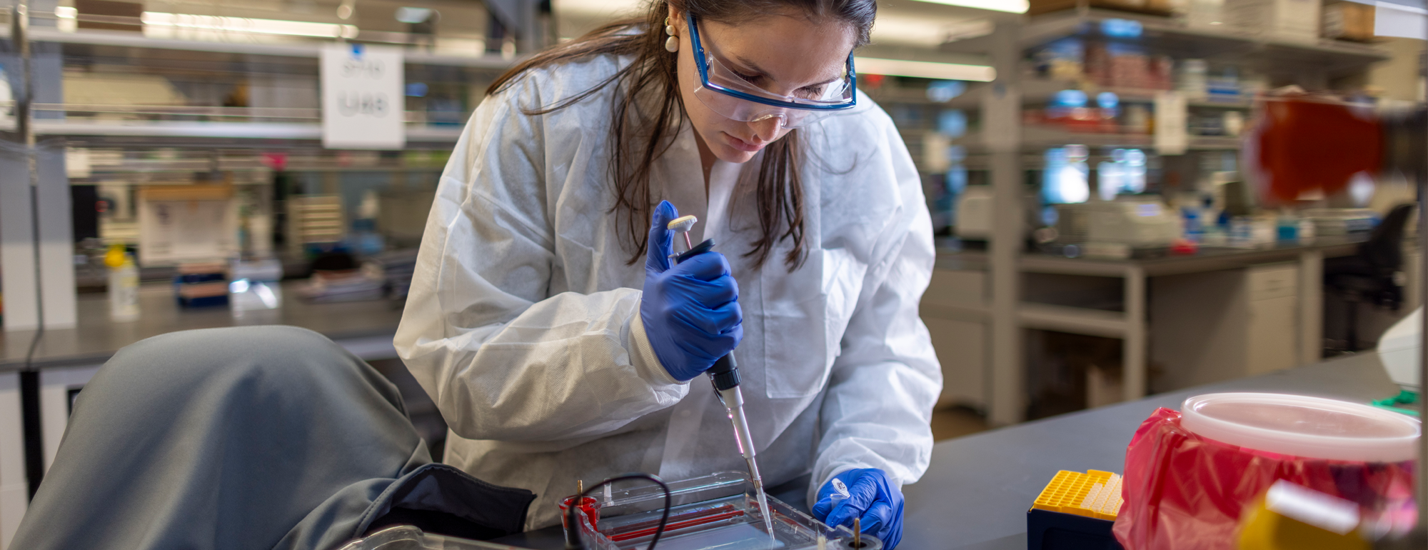 A resident in a lab wearing a white coat, gloves and goggles is pipetting.