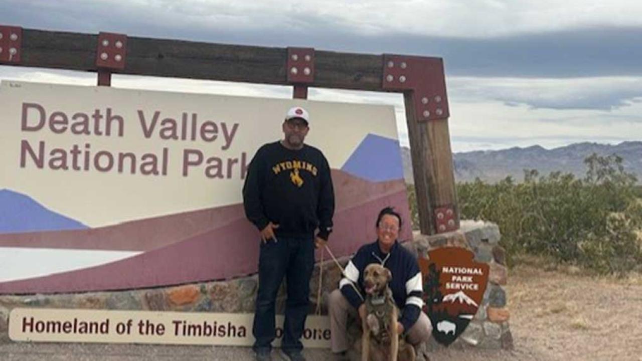 A man, a woman, and dog in front of the Death Valley National Park sign with mountains behind.