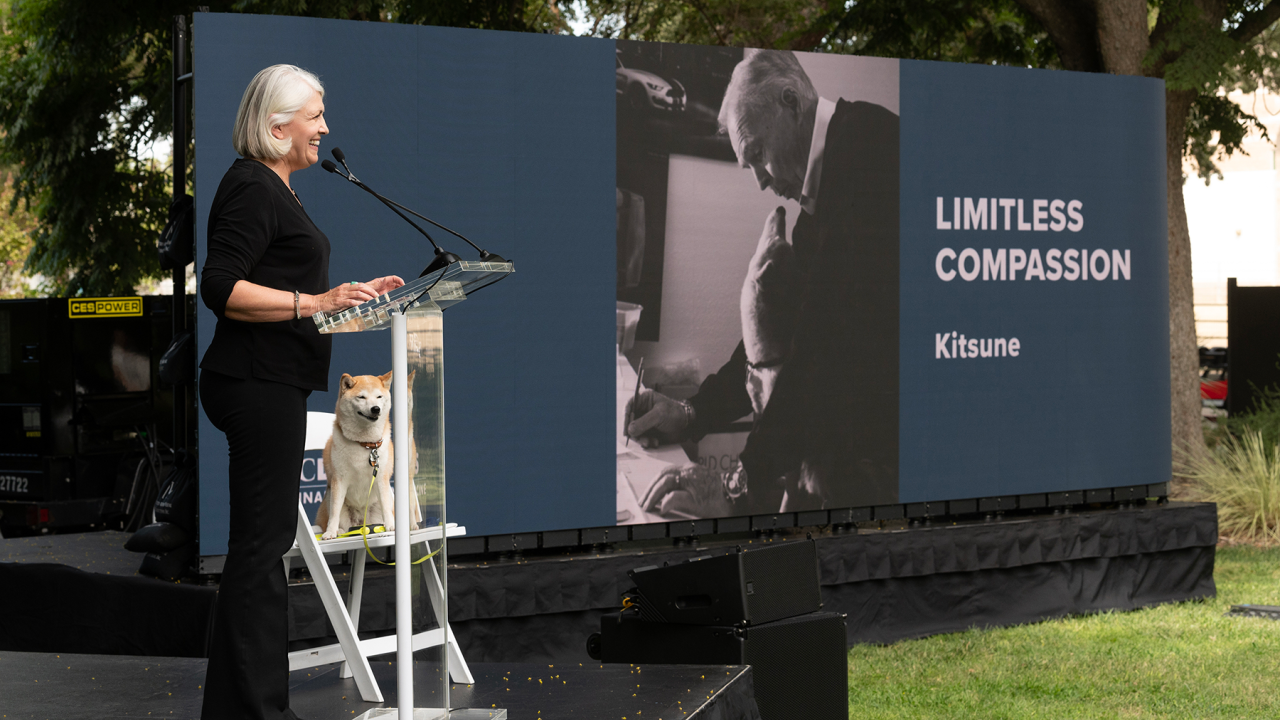 A person standing at a podium on an outdoor stage with a dog sitting on the stage in a chair and a monitor in the background with the words "limitless compassion."