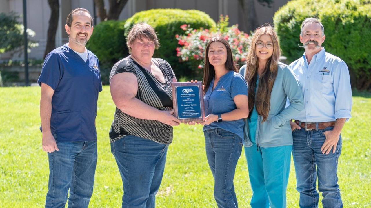 Six adults on lawn smiling, holding an award plaque in a sunny park
