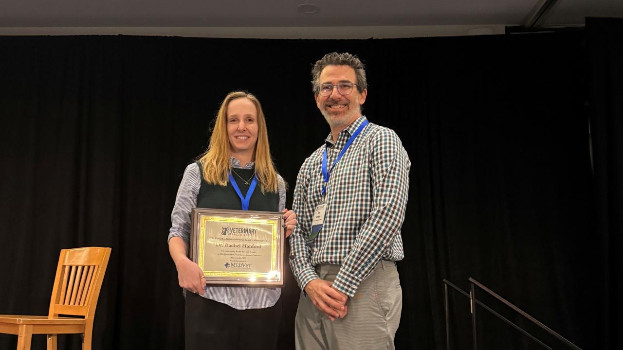 Two smiling people on stage holding a framed award plaque