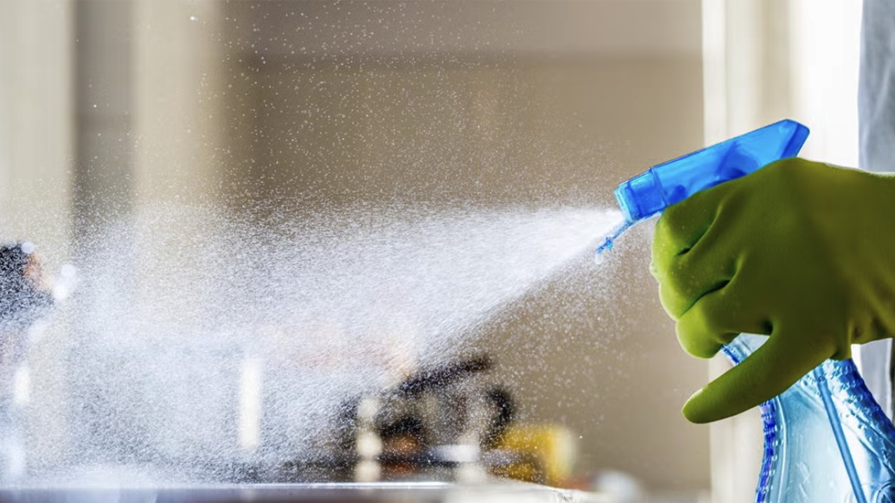 A gloved hand sprays cleaning solution, creating a mist in a bright kitchen.