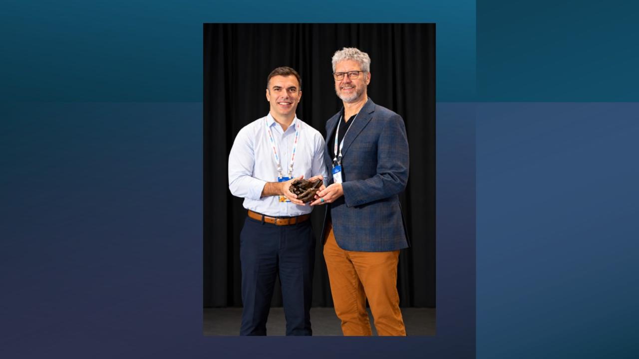 Two men in smart casual attire smiling on stage; younger man holds an award