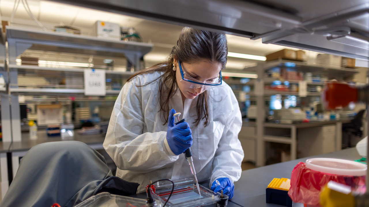 A lab technician dressed in a white coat, wearing gloves and googles is pipetting.