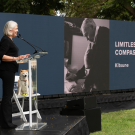 A person standing at a podium on an outdoor stage with a dog sitting on the stage in a chair and a monitor in the background with the words "limitless compassion."