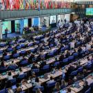Conference hall with rows of delegates at desks, national flags draped along the upper gallery