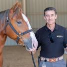 Smiling man in black shirt in indoor arena holding lead rope beside chestnut horse