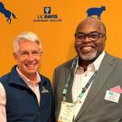 Two smiling men pose together in front of an orange backdrop with animal logos and text.